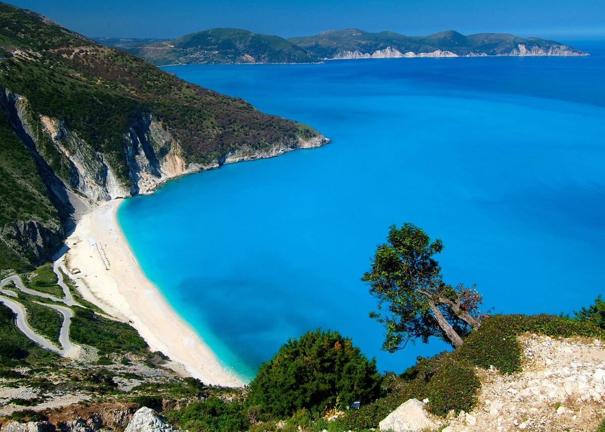 A serene beach is depicted, bordered by steep green hills and cliffs. The clear blue waters gently lap at the sandy shore, creating a tranquil setting. The coastline curves gracefully, inviting exploration of the surrounding natural beauty.