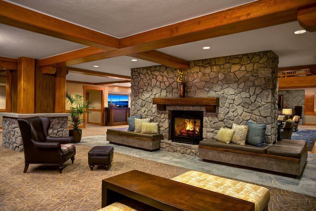 A spacious lobby is highlighted by a stone fireplace set within a rustic stone wall, complemented by wooden beams overhead. Comfortable seating options include plush chairs and benches adorned with decorative cushions. Soft lighting creates a cozy ambiance, inviting guests to relax.
