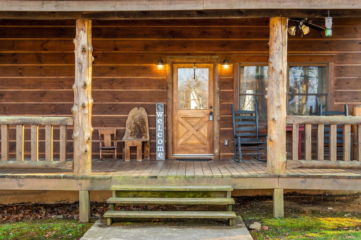The cabin's welcoming front porch features natural wood accents and a stone walkway leading to the door. Two rocking chairs and a rustic bench add to the inviting atmosphere, complemented by a 'Welcome' sign placed prominently. Warm lighting enhances the entryway.