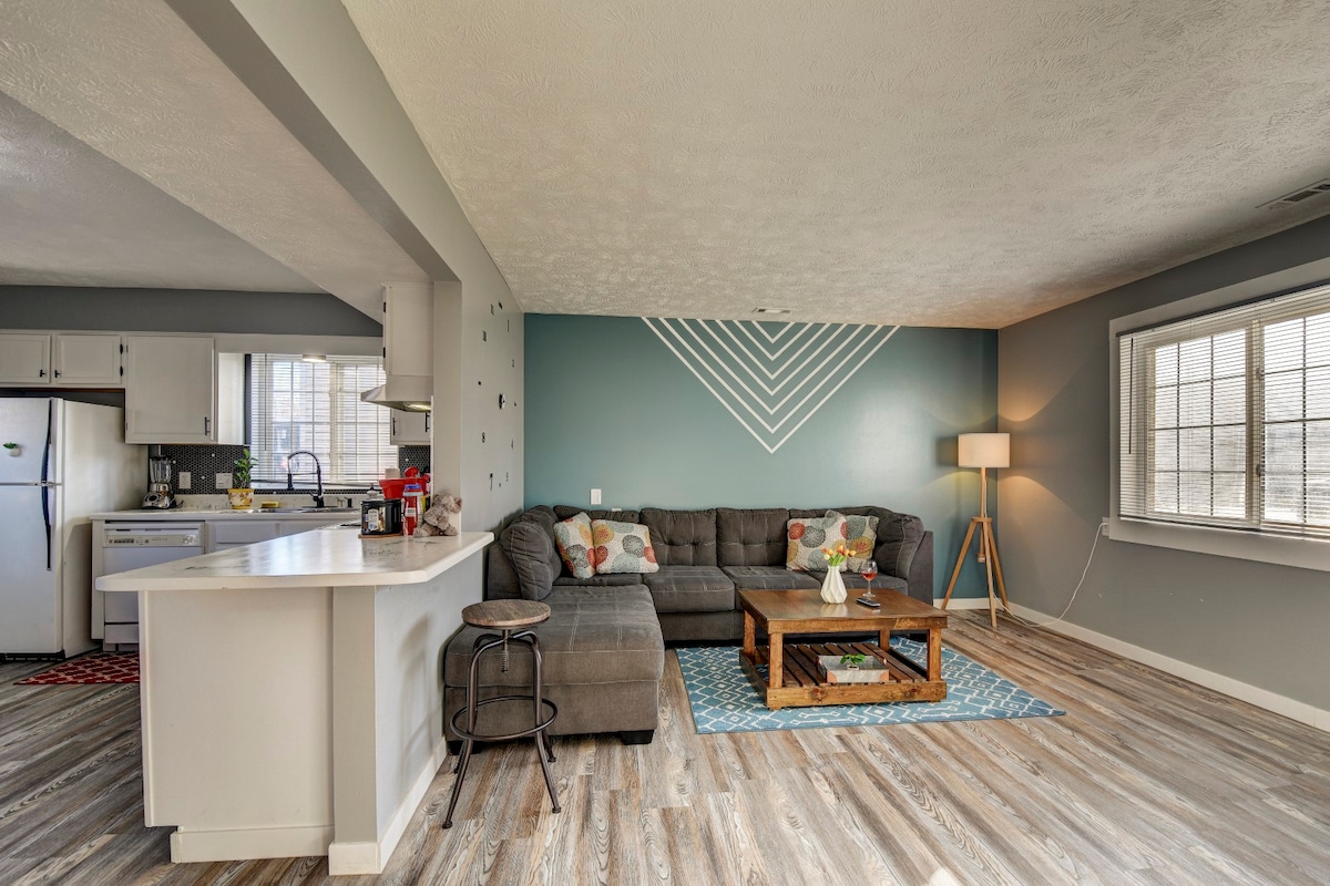 A spacious living area features a sectional couch in a neutral tone, paired with a wooden coffee table. A floor lamp adds soft lighting, while a patterned area rug enhances the space. The adjacent kitchen, visible in the background, is equipped with modern appliances.