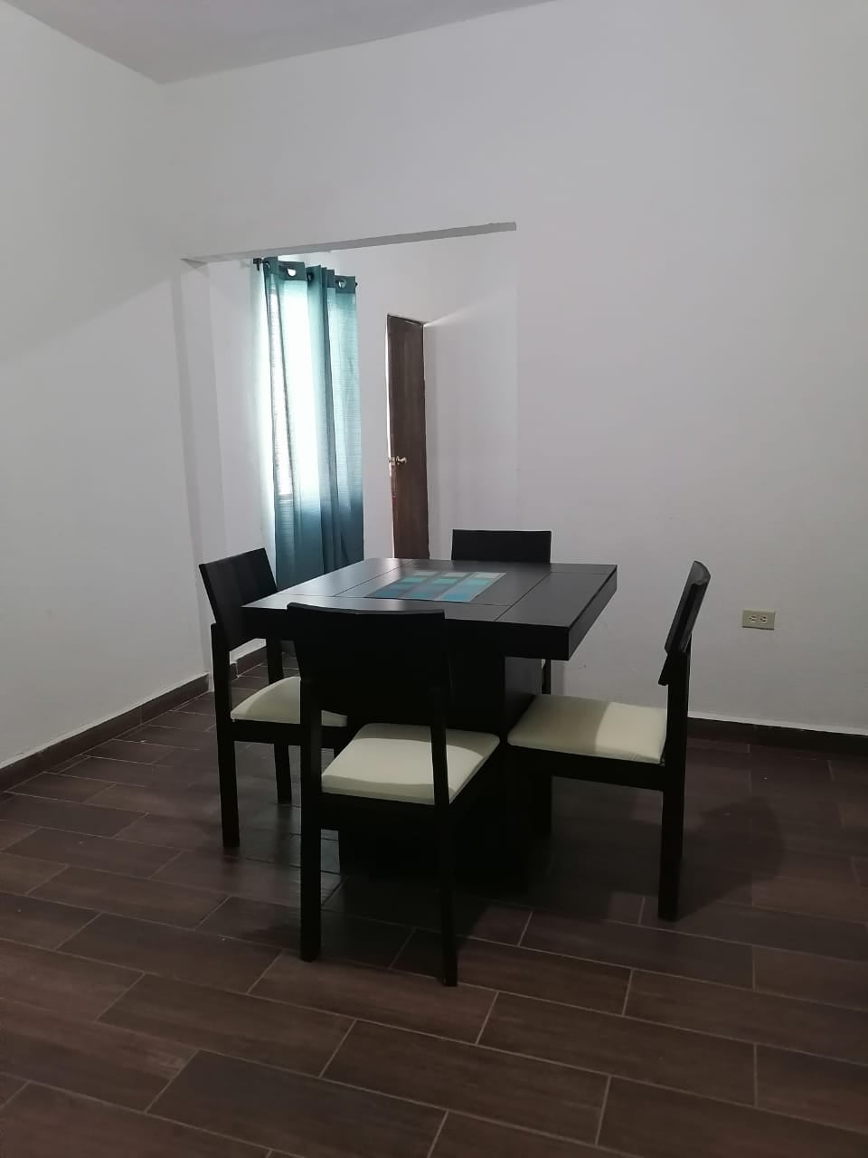 A dining area is presented with a square black table surrounded by four chairs. Light-colored cushions accentuate the chairs. A window with sheer teal curtains allows natural light to enter, and a wooden door is visible in the background, enhancing accessibility.