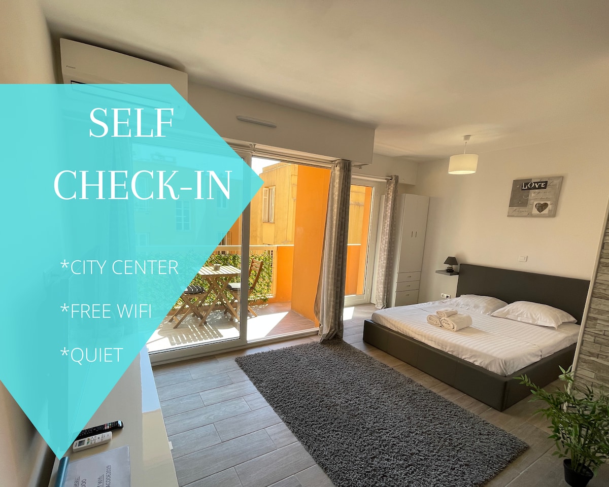 The studio features a modern sleeping area with a spacious bed placed against a light-colored wall. Large glass doors open onto a terrace, allowing natural light to fill the room. The area is complemented by a cozy rug and a wardrobe for storage.