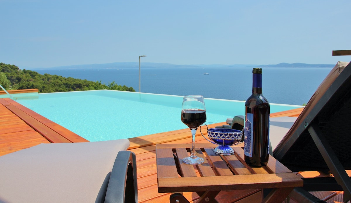 A spacious wooden deck features an infinity pool overlooking the sea, with a glass of red wine and a bottle on a small table. Comfortable lounge chairs are positioned nearby, providing an inviting spot to relax and enjoy the tranquil view of the water and distant coastline.