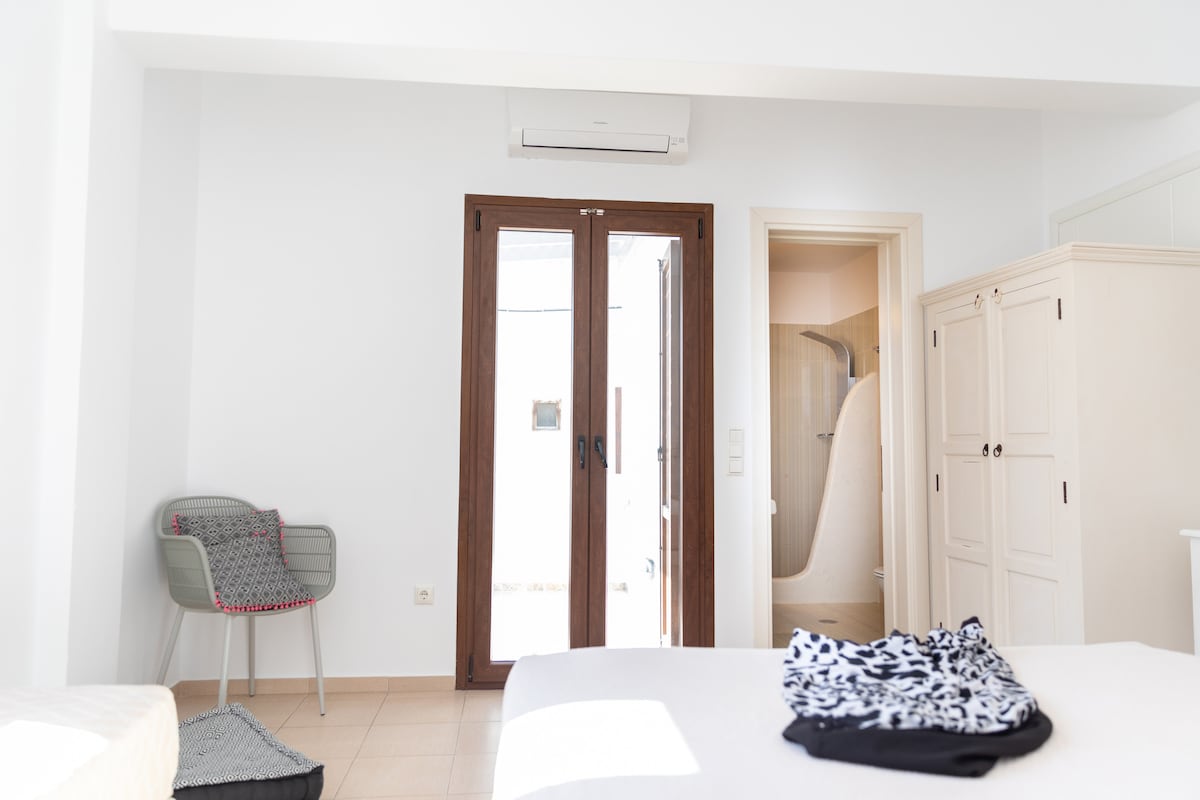 A bright room features large double doors leading to an outdoor space. A comfortable chair sits in the corner, and a neatly made bed is positioned in view. Light-colored walls create a spacious feel, complemented by light flooring.