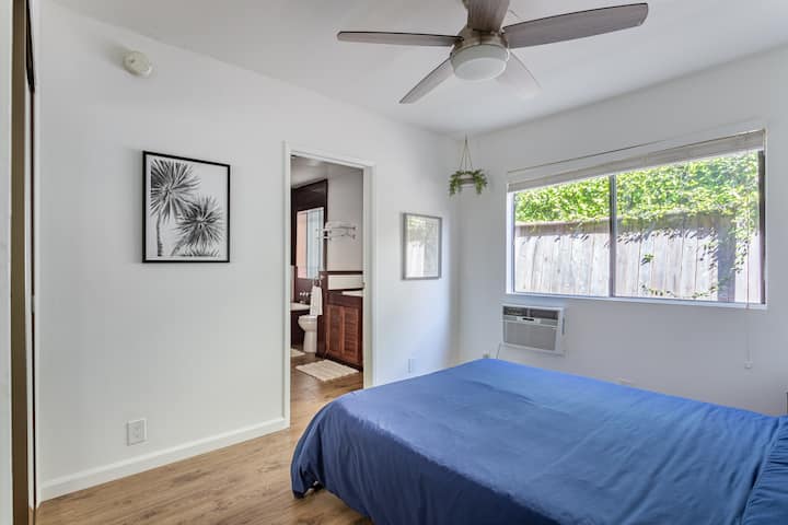 Un dormitorio minimalista con ventilador de techo, colcha azul y aire acondicionado. Una gran ventana revela la vegetación al aire libre. La habitación cuenta con paredes ligeras, suelos de madera y vistas a un baño adyacente. Bañera hundida