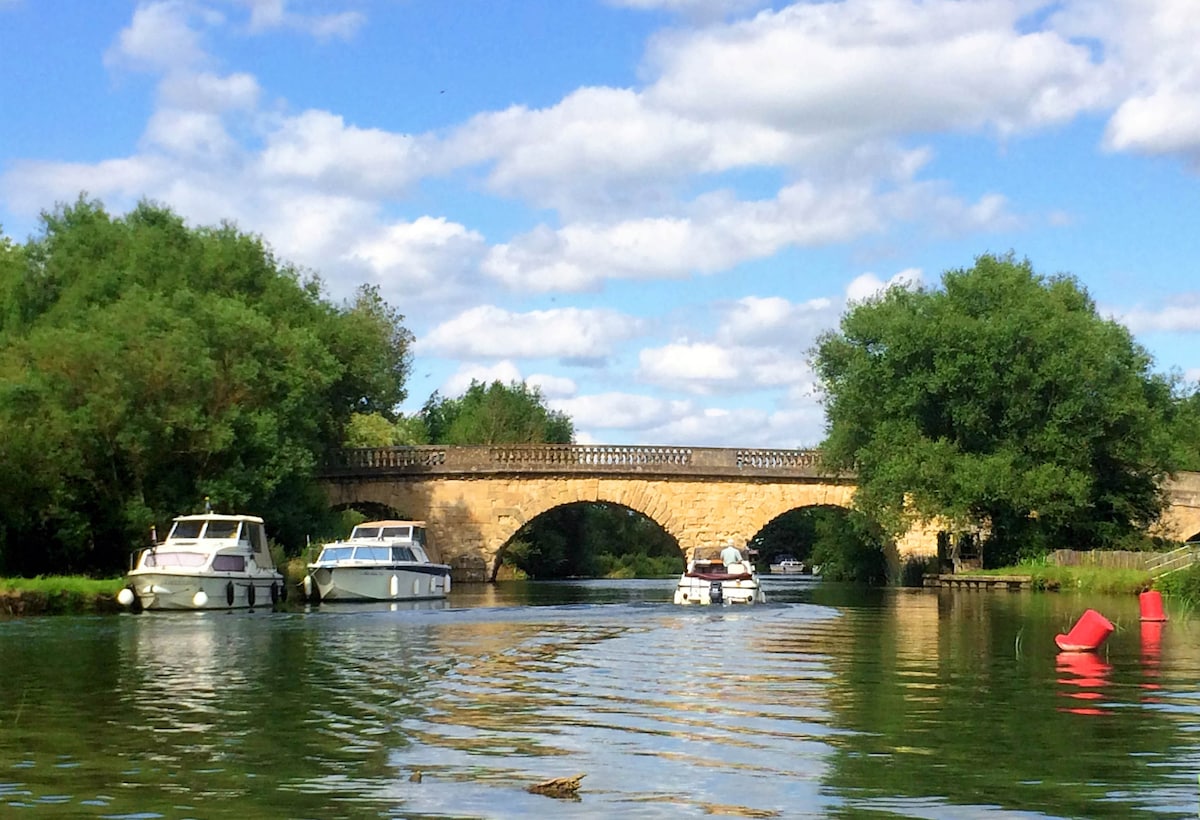 A scenic view of a stone bridge arches over a shimmering river, flanked by lush green trees. Several boats are moored along the banks, while the sky above is filled with fluffy clouds, reflecting a peaceful and natural setting.