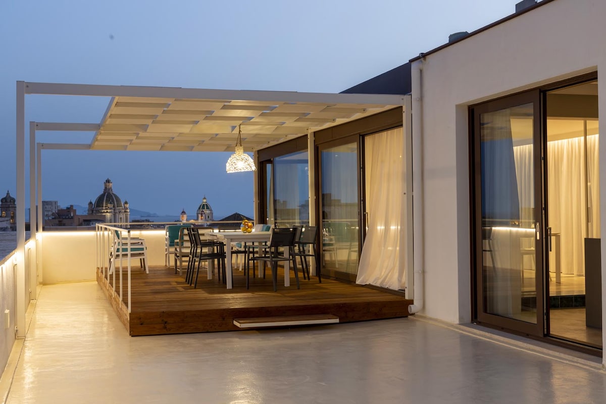 An outdoor terrace features a modern dining set with multiple chairs, complemented by a large umbrella for shade. Glass doors provide access to the interior, showcasing softly lit curtains. The surrounding view includes city rooftops, highlighting architectural details in the evening light.