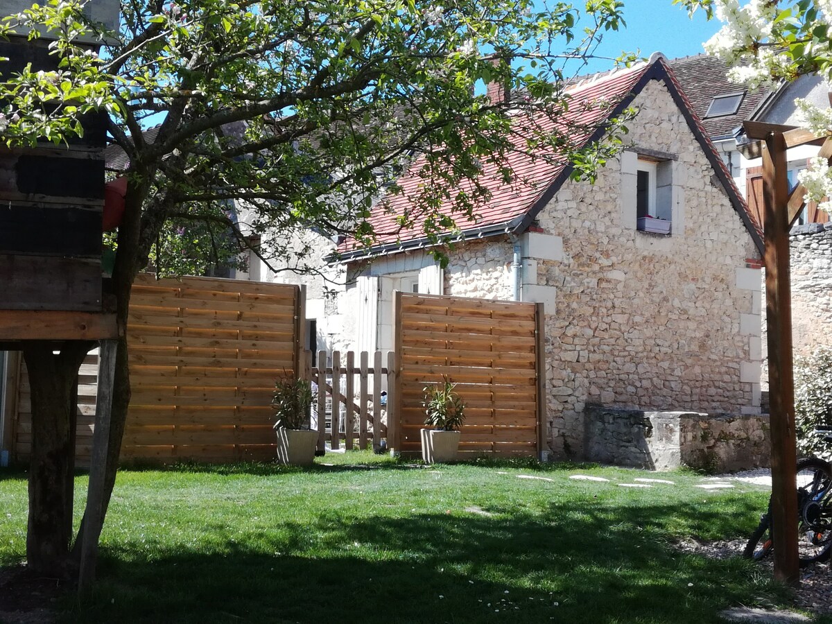 A charming stone house is framed by a green lawn, featuring wooden fences that provide privacy. The building showcases a classic roof and is complemented by blooming trees in the foreground, set against a bright blue sky.