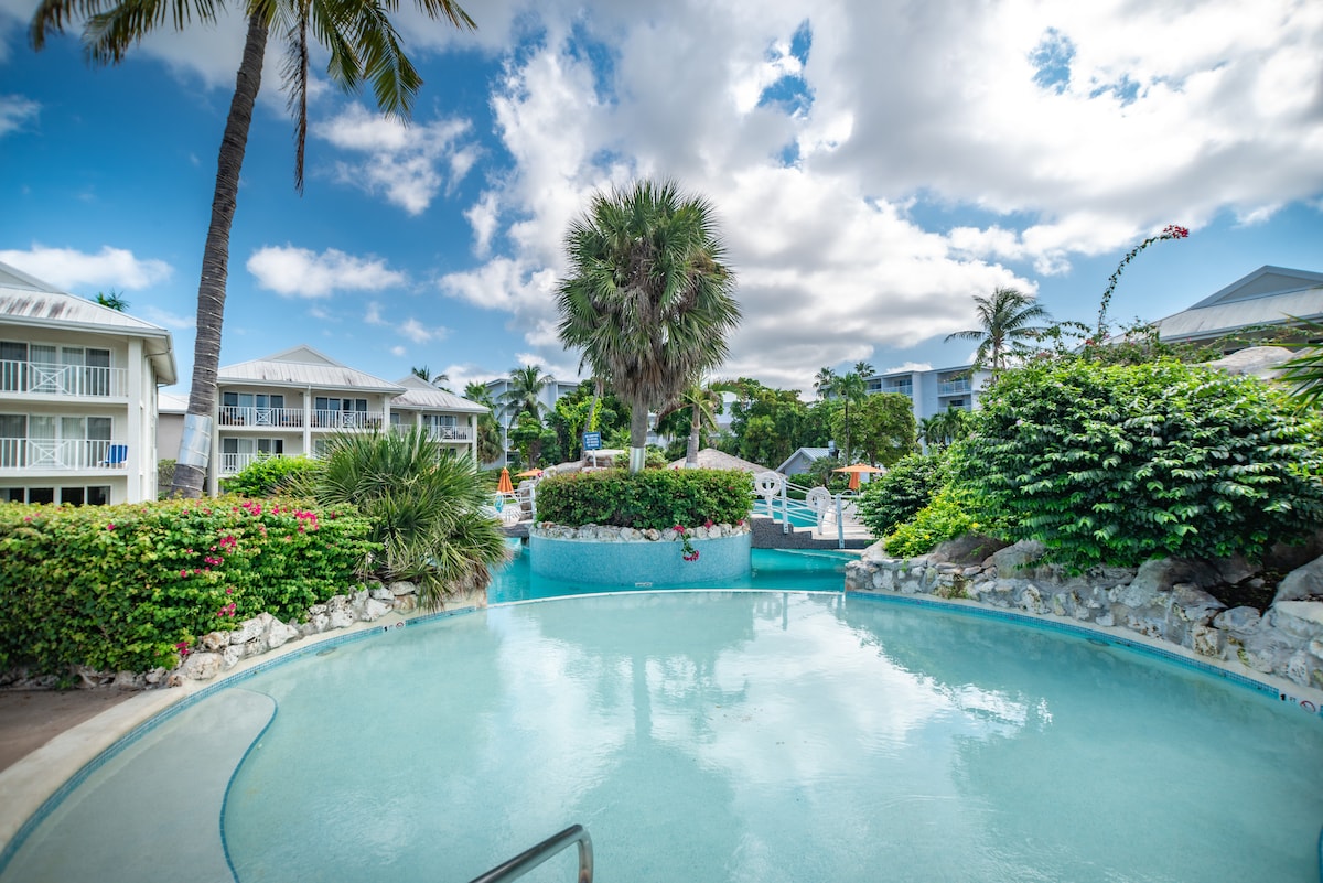 The inviting pool area features a winding turquoise pool surrounded by lush tropical landscaping. Palm trees provide shade, while the bright sky adds a serene ambiance. Lounge chairs are situated on the deck for relaxation, complemented by colorful flowering plants throughout the area.