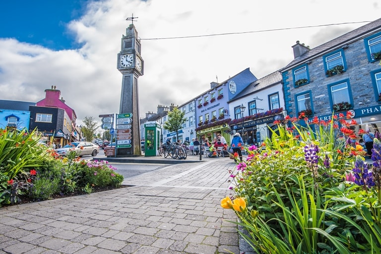 A vibrant town square is depicted, featuring a tall clock tower surrounded by lush flowerbeds blooming with various colors. Shops with charming facades line the street, while bicycles are parked near the entrance, and pedestrians enjoy a leisurely stroll through the area.