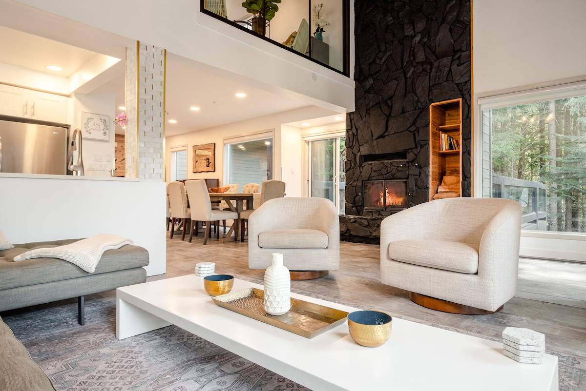 A cozy living area features two rounded armchairs facing a central coffee table with decorative items. A stone fireplace adds warmth to the space, while large windows provide natural light and views of the surrounding greenery.