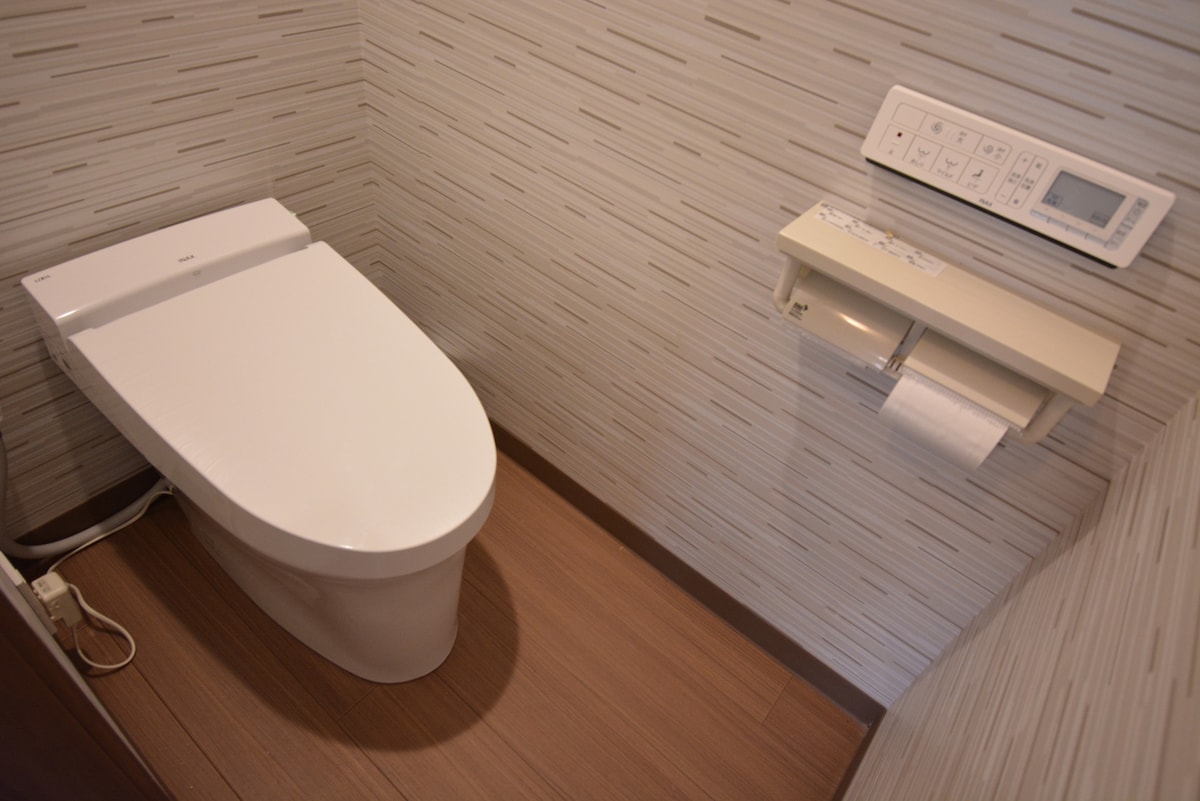 A modern bathroom features a sleek, white toilet positioned on a wooden floor. A control panel is mounted on the wall nearby, alongside a toilet paper holder, enhancing accessibility and convenience. Neutral-colored wall coverings add to the clean aesthetic of the space.