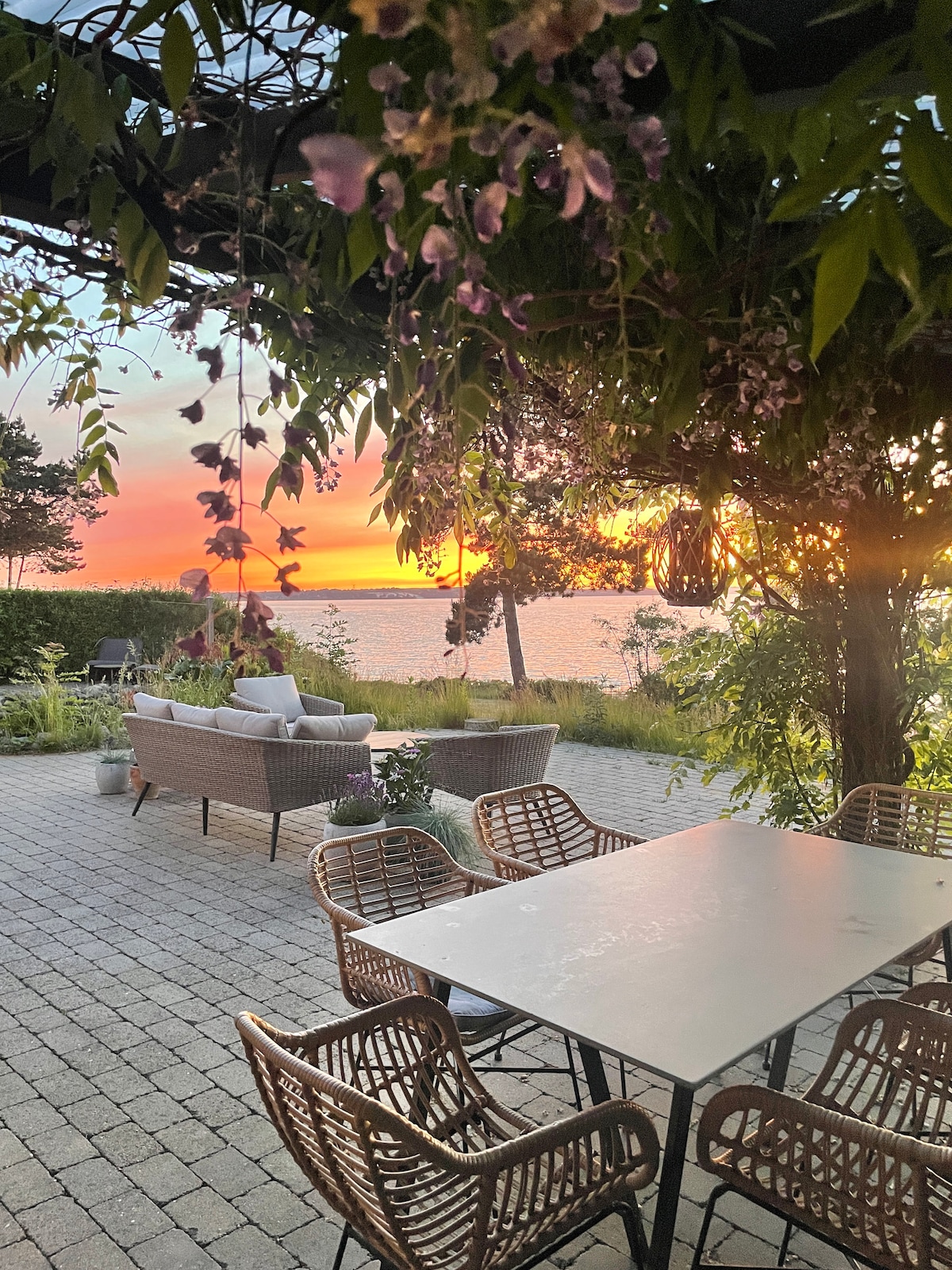 An outdoor seating area is showcased with a table surrounded by woven chairs. In the background, a sunset casts vibrant colors over the water, while lush greenery and flowers frame the scene, enhancing the tranquil atmosphere.