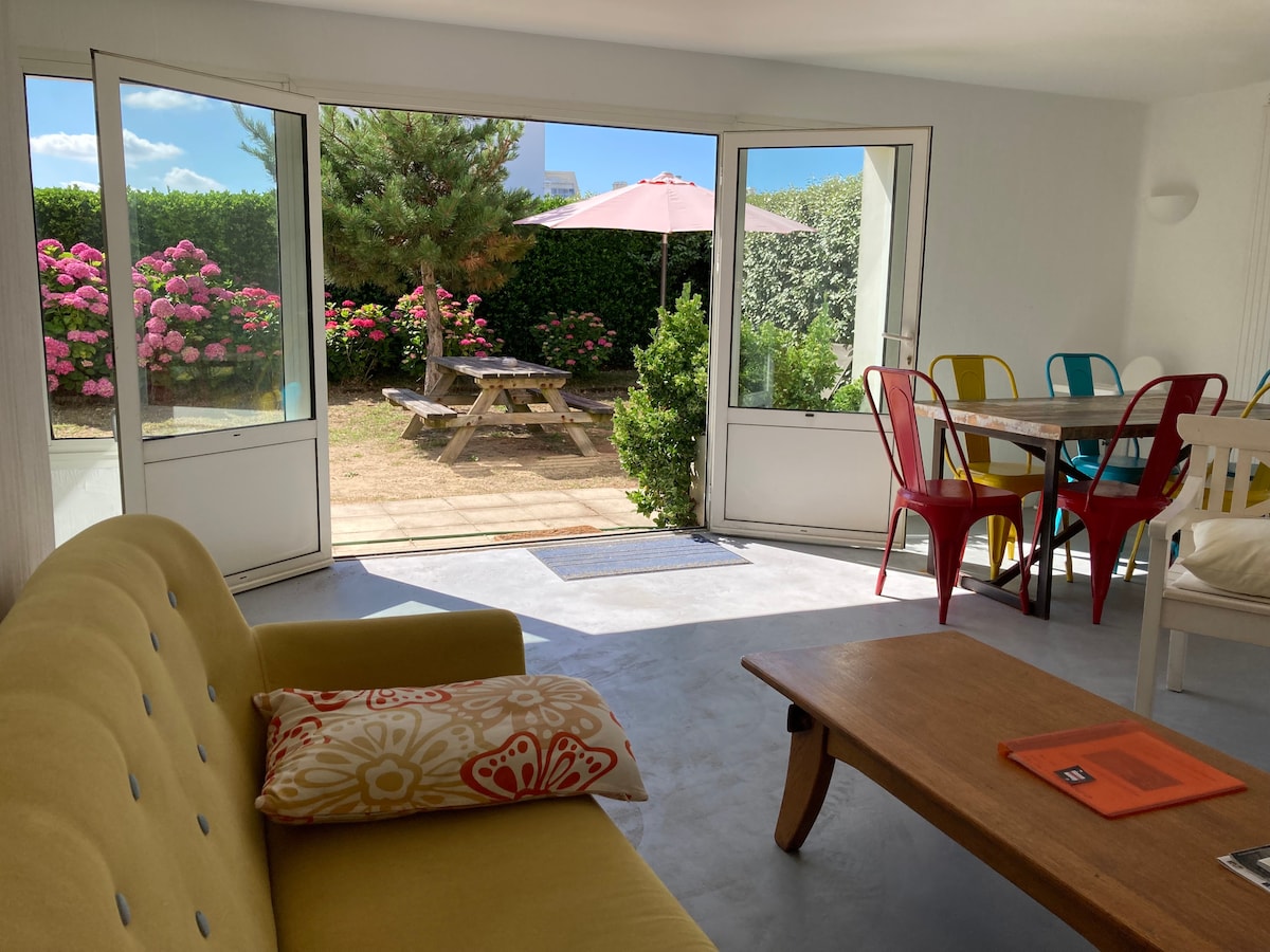 A bright living area is showcased with large glass doors, opening to a garden scene filled with vibrant pink flowers. Colorful chairs surround a dining table, while a yellow sofa with a decorative pillow offers seating. Sunlight enters through the open doors, highlighting the indoor space.