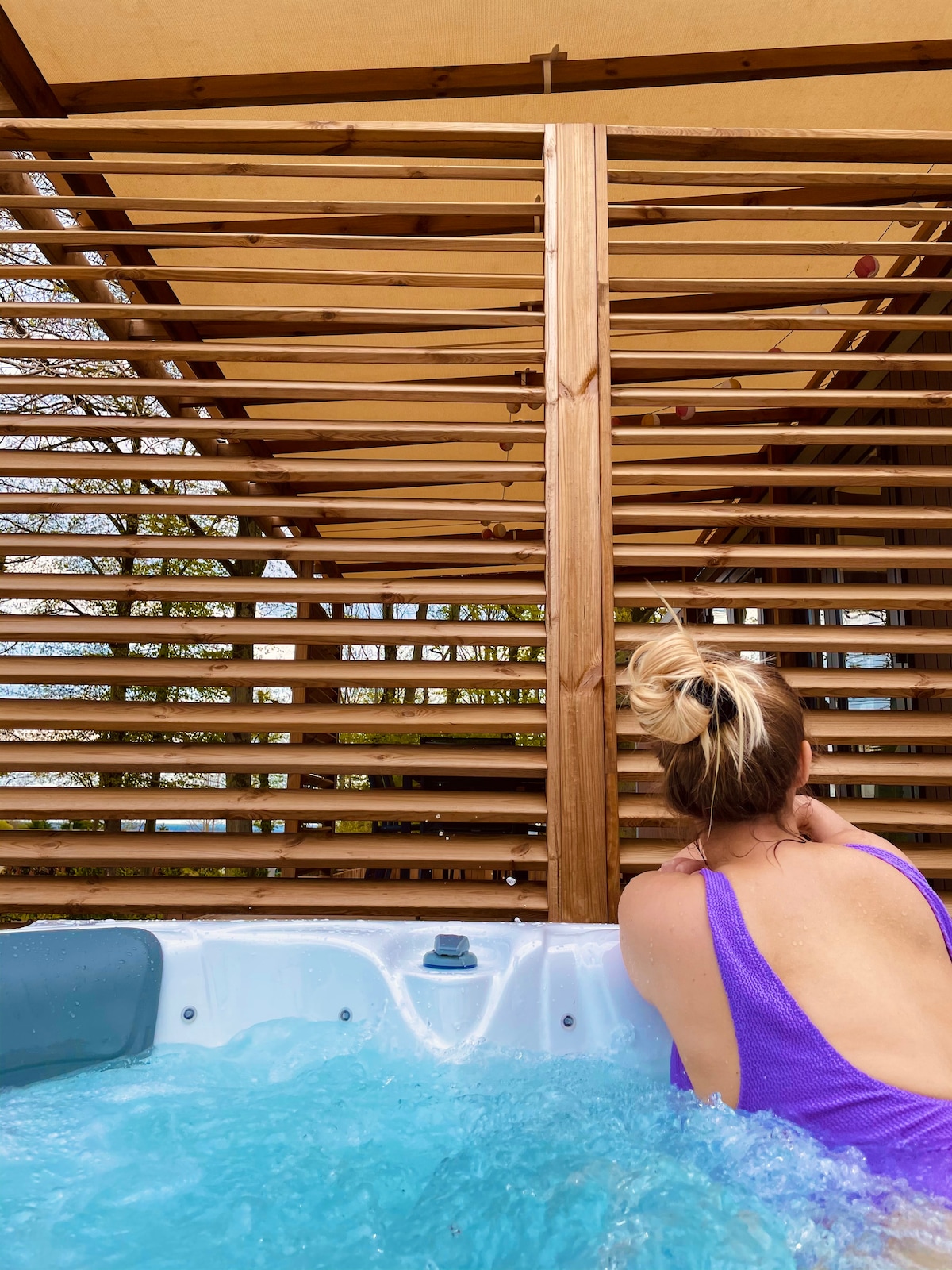 A private jacuzzi is surrounded by wooden slatted walls, providing both privacy and a connection to nature. The inviting water is illuminated, while a person relaxes, facing away from the camera. Natural light filters through the slats, enhancing the serene environment.