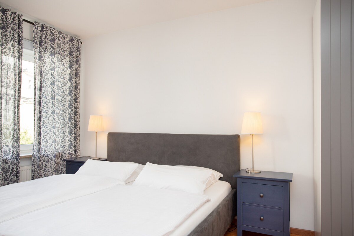 A cozy sleeping area features a spacious bed with crisp linens. Two bedside tables with lamps provide soft lighting. Floor-to-ceiling windows are adorned with patterned curtains, allowing natural light to fill the room.