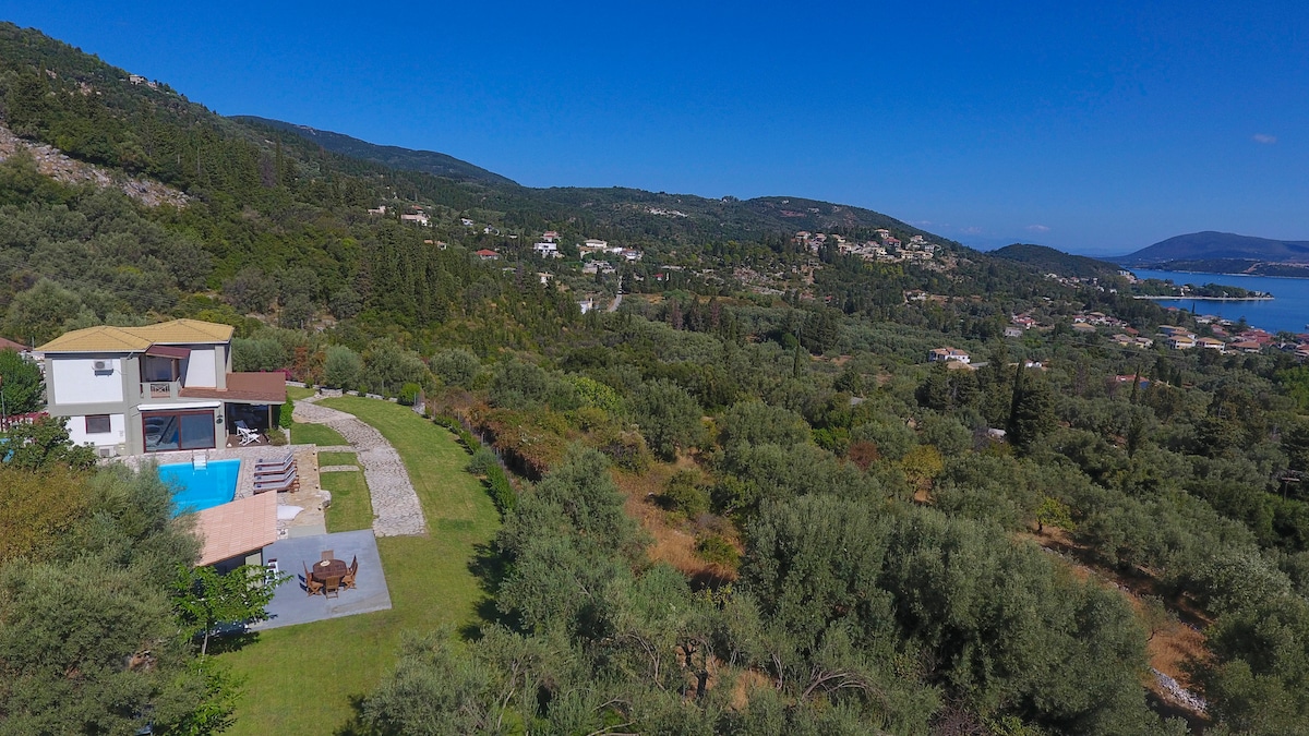 An aerial view of Villa Artemis is presented, showcasing its large garden area and private pool. Olive trees and lush greenery surround the property, enhancing the scenic landscape. The villa's modern structure is visible, nestled within a quiet and peaceful environment.