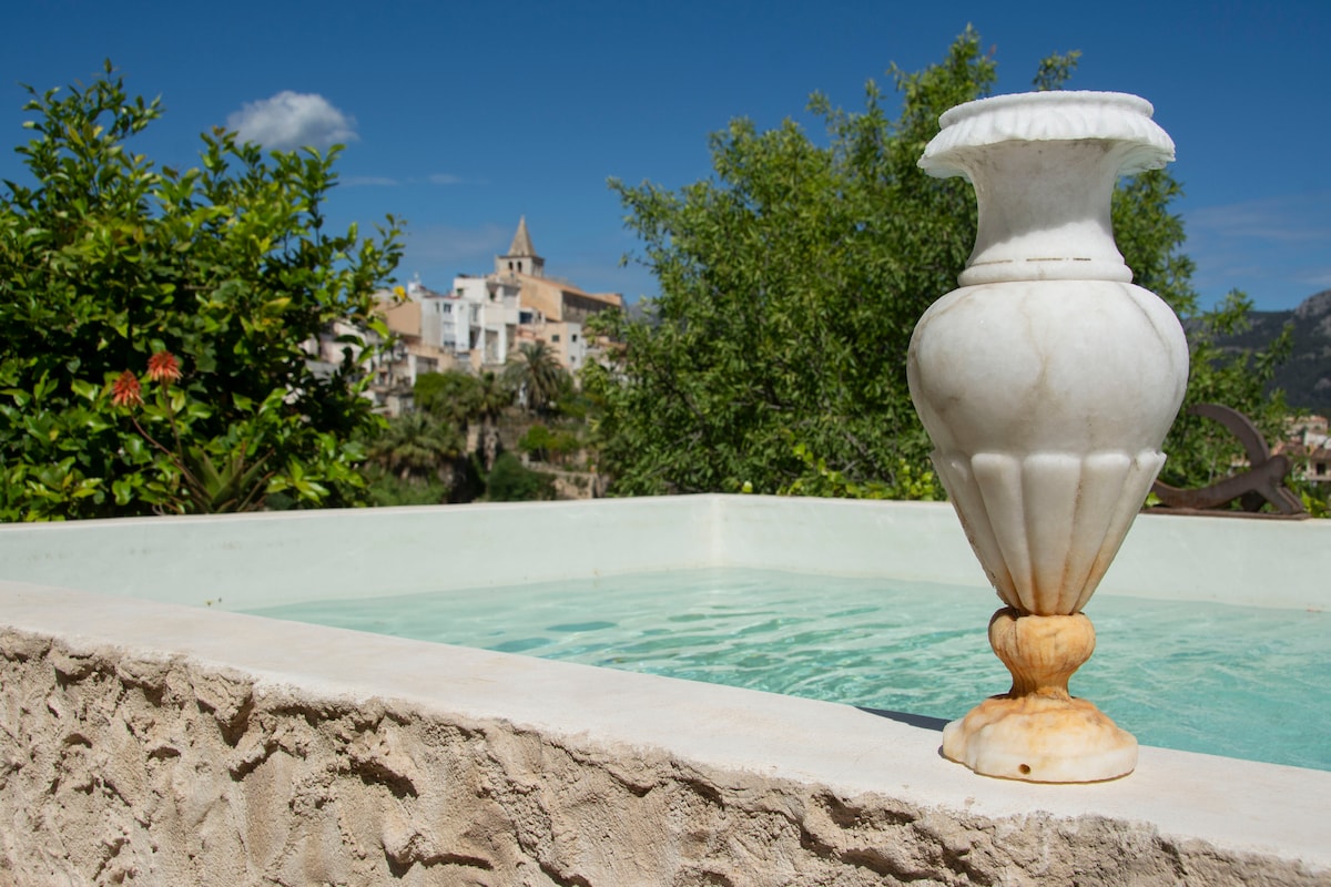 A marble vase stands elegantly at the edge of a tranquil water feature, with crystal clear water reflecting the sky. In the background, a charming village nestled in the hills is visible, framed by greenery and under a blue sky.