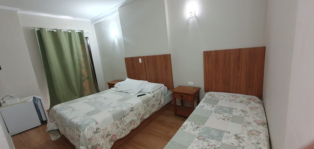 A room features two beds with floral-patterned bedding, accompanied by wooden nightstands on either side. Soft lighting enhances the neutral-toned walls, and a green curtain partially covers a window, providing a touch of color to the serene environment.