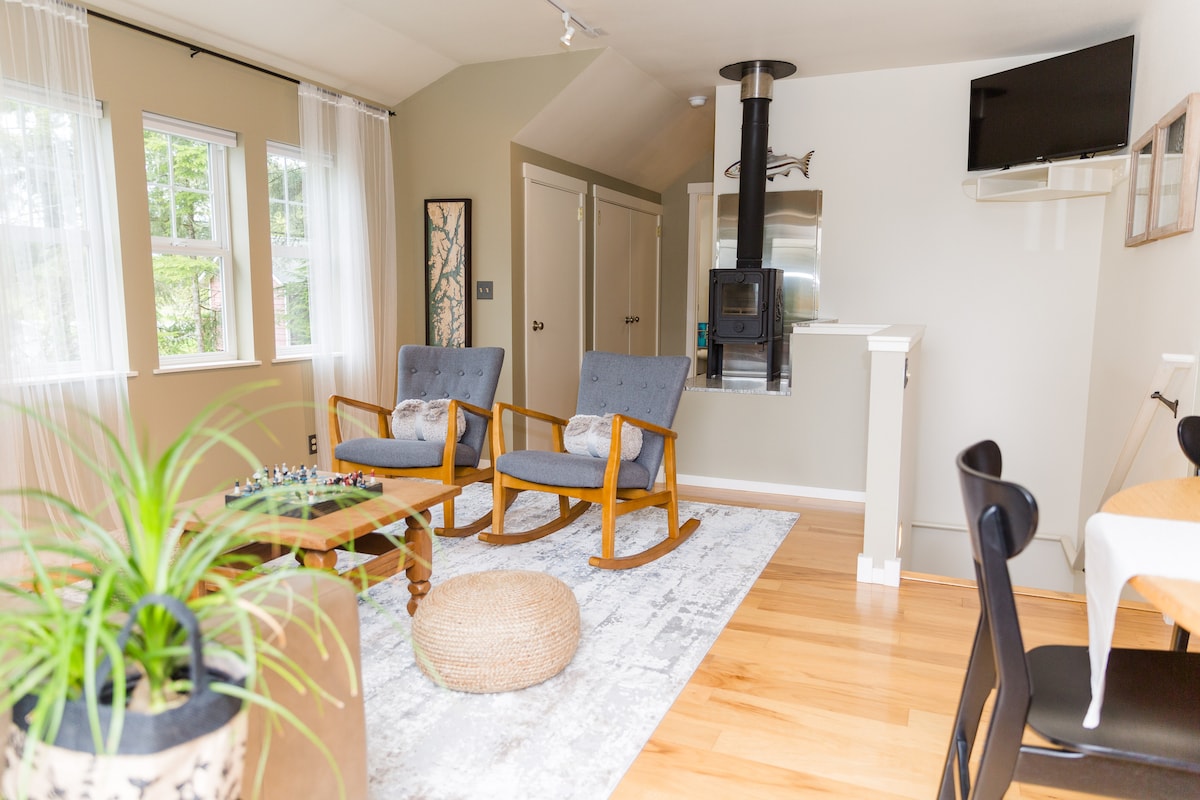 A bright and welcoming living area features two rocking chairs positioned around a wooden coffee table. A cozy area rug lies on the hardwood floor, complemented by a pouf nearby. Large windows allow natural light to fill the space, highlighting the warmth of the interior.