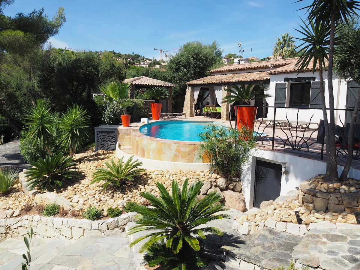 The image showcases a private swimming pool with a stone surround, surrounded by lush Mediterranean vegetation and decorative rocks. Vibrant orange planters add a pop of color, while a charming villa can be seen in the background, framed by trees and greenery.