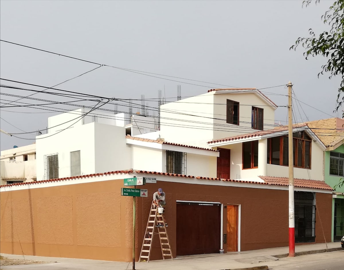 A two-story European-style house is displayed, featuring white walls and a tiled roof. Large windows allow natural light to enter, while a wooden door provides access. A ladder leans against the house, indicating ongoing maintenance. Power lines and neighboring buildings are visible in the background.
