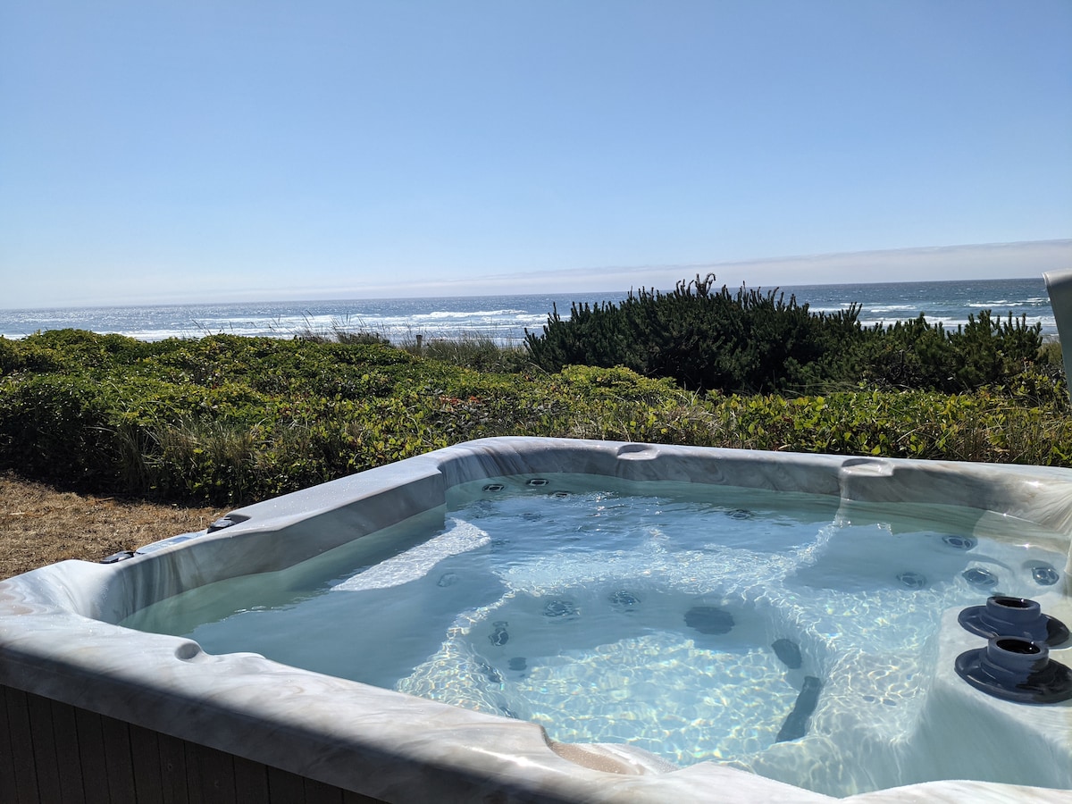 A hot tub is positioned outdoors, offering expansive views of the ocean and lush greenery. Gentle waves of the water can be seen in the distance, under a clear blue sky. The tub’s smooth edges and inviting surface contrast with the natural surroundings.