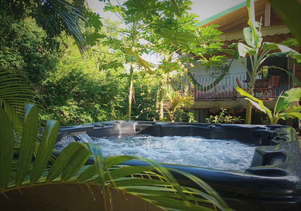 A private jacuzzi is situated among lush greenery, surrounded by tropical plants. Bubbling water offers a feeling of relaxation, while a bungalow is visible in the background, featuring a hammock and seating area on the porch, set against a serene, natural backdrop.