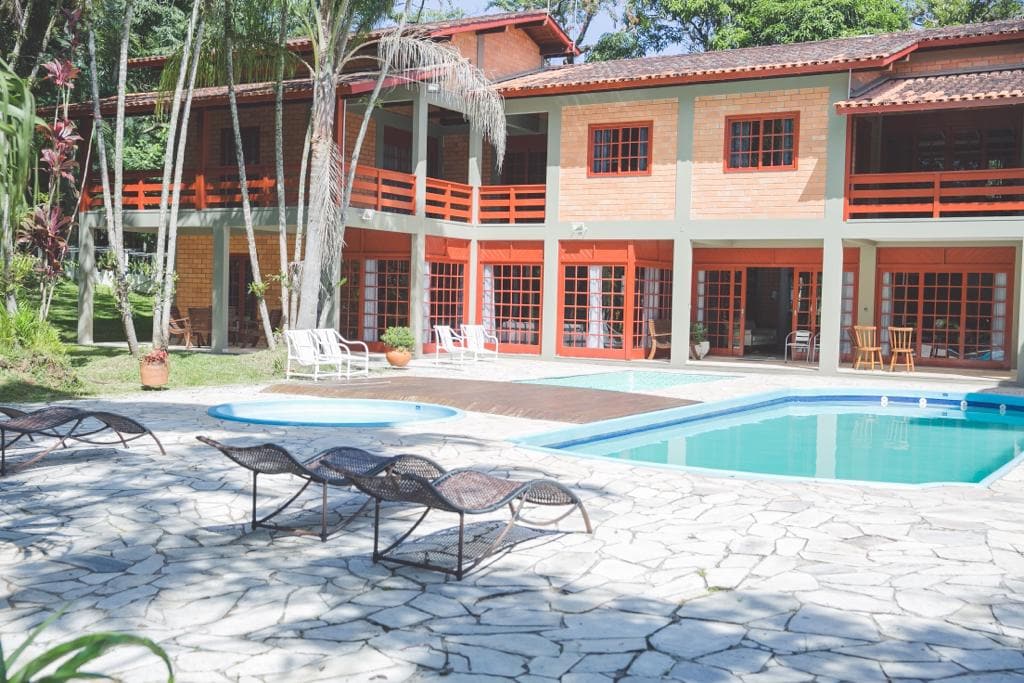 A spacious outdoor area features two swimming pools surrounded by a stone patio. Comfortable lounge chairs are positioned under the sun, and a multi-level house with large windows and red accents is visible in the background, enhancing the inviting atmosphere.