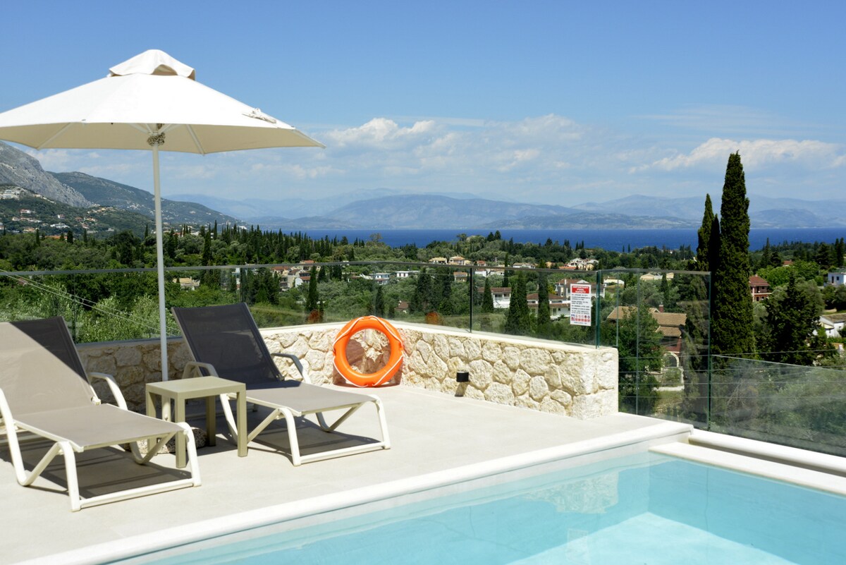 A poolside view showcases two lounge chairs under a sun umbrella, positioned along the edge of a modern infinity pool. The landscape offers a sweeping view of rolling hills and the coastline, with varying shades of green and blue under a clear sky.