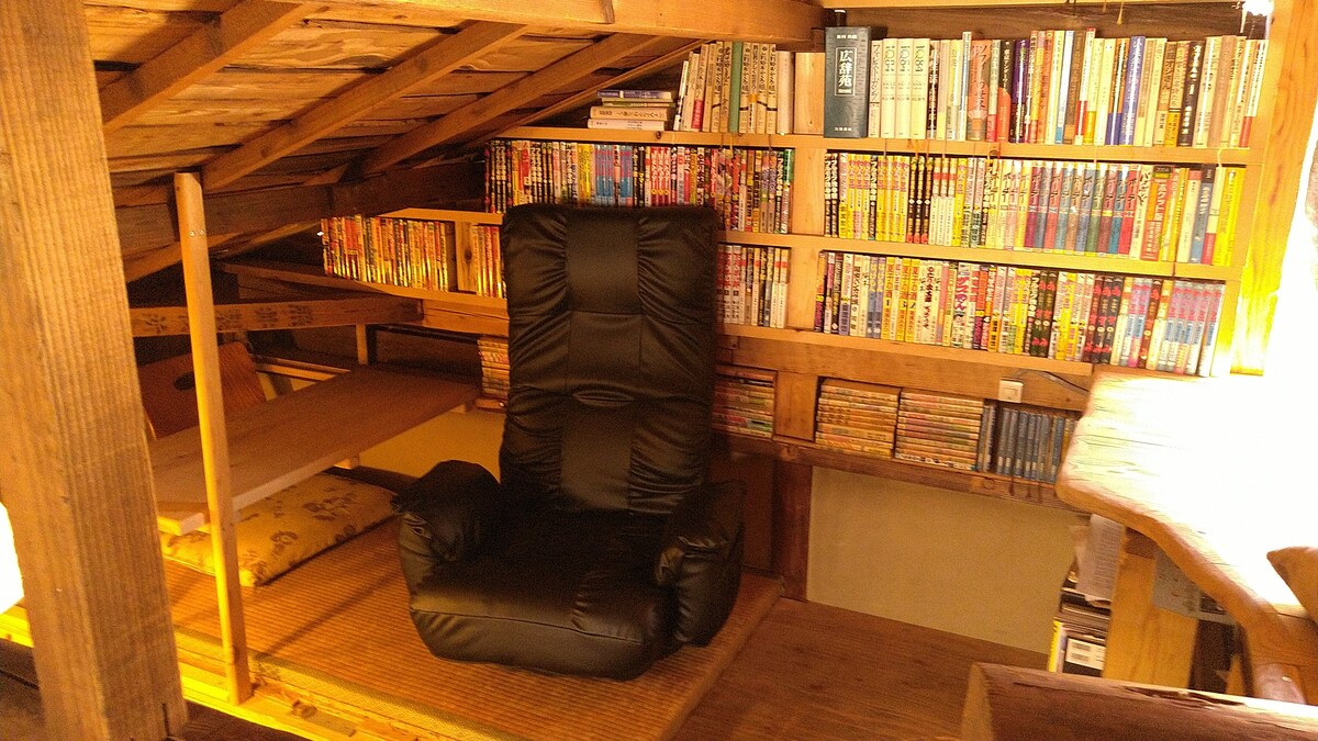 A cozy nook features a black reclining chair positioned next to a wall lined with colorful bookshelves filled with various comic books and novels. A soft mat is accessible nearby, providing additional seating or resting space.