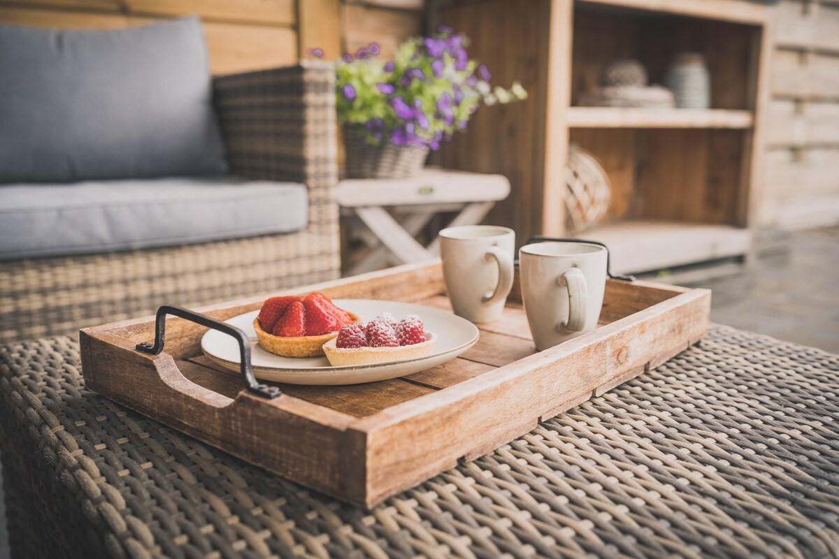 A wooden tray is positioned on a textured table, featuring fresh strawberries and a pastry. Two ceramic mugs are placed nearby, complementing the inviting setting. The background includes soft seating and decorative items, contributing to a relaxed atmosphere.