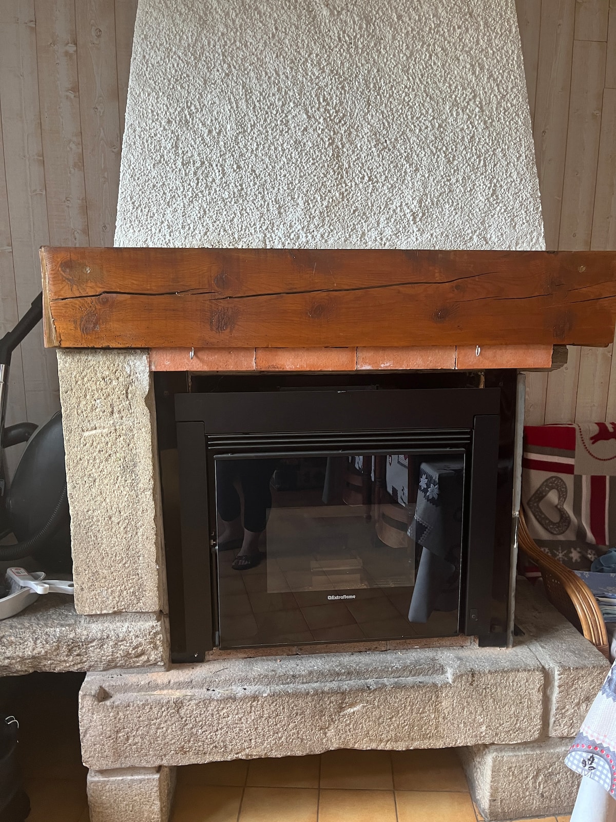 A modern pellet stove is set within a rustic stone fireplace, featuring a charming wooden mantel above. The stove's glass front reflects the surrounding space. The warm tones of the stone and wood create a welcoming focal point within the room.
