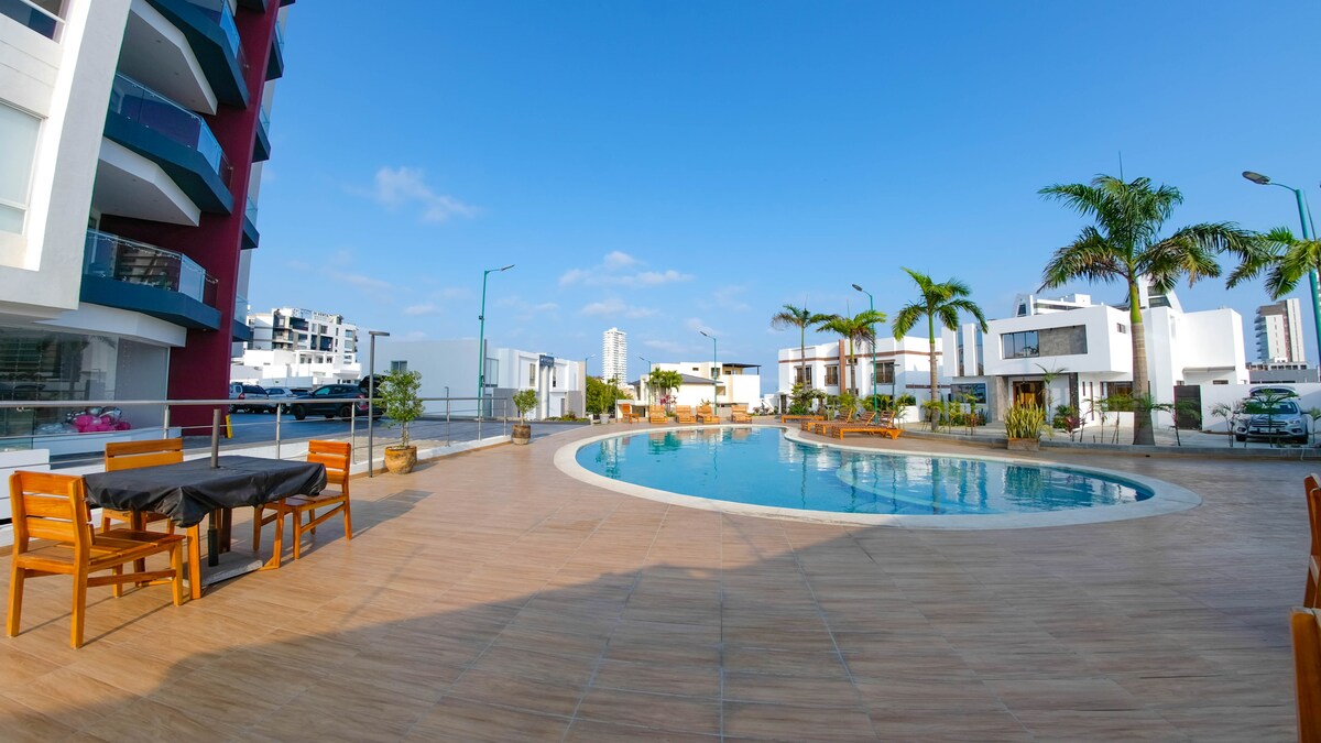 A spacious outdoor area features a curved swimming pool surrounded by palm trees and lounge chairs. Dining tables are arranged nearby, offering a view of the water. The surrounding architecture contributes to an open atmosphere under a clear blue sky.