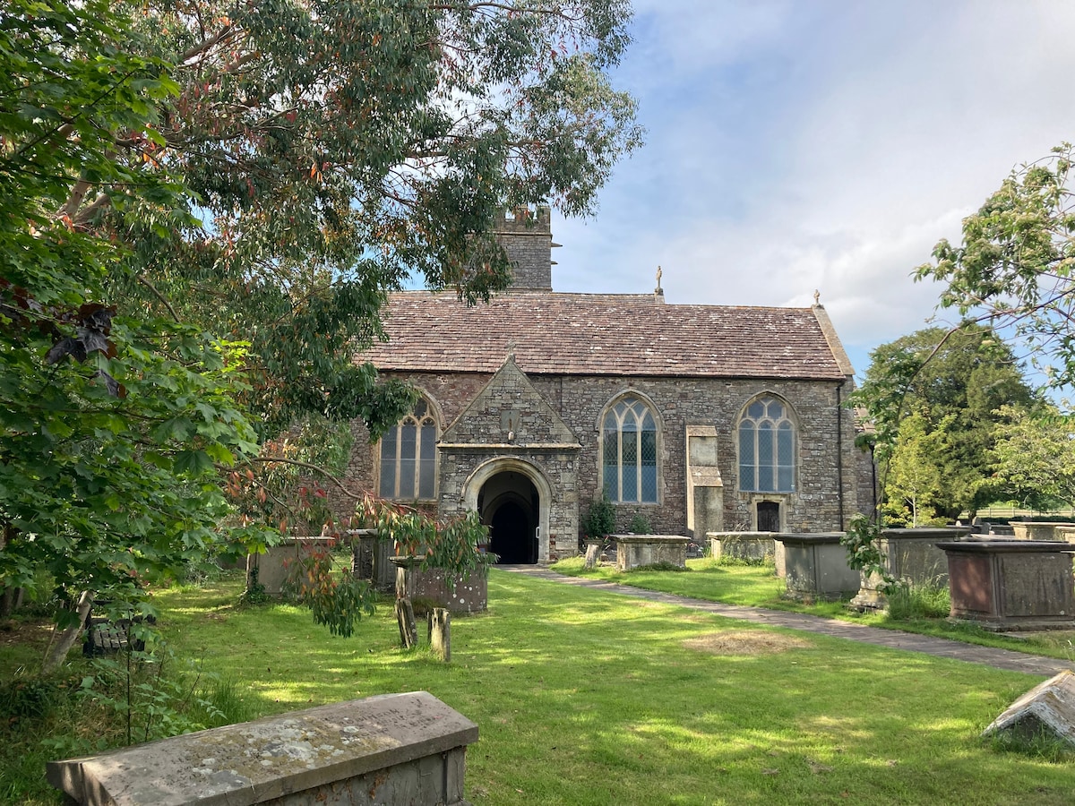 An ancient stone church stands amidst greenery, featuring a slate roof and tall arched windows. Surrounding the building are gravestones, set on a well-maintained lawn, with trees providing a natural frame. The structure is enhanced by a peaceful atmosphere, inviting moments of reflection.