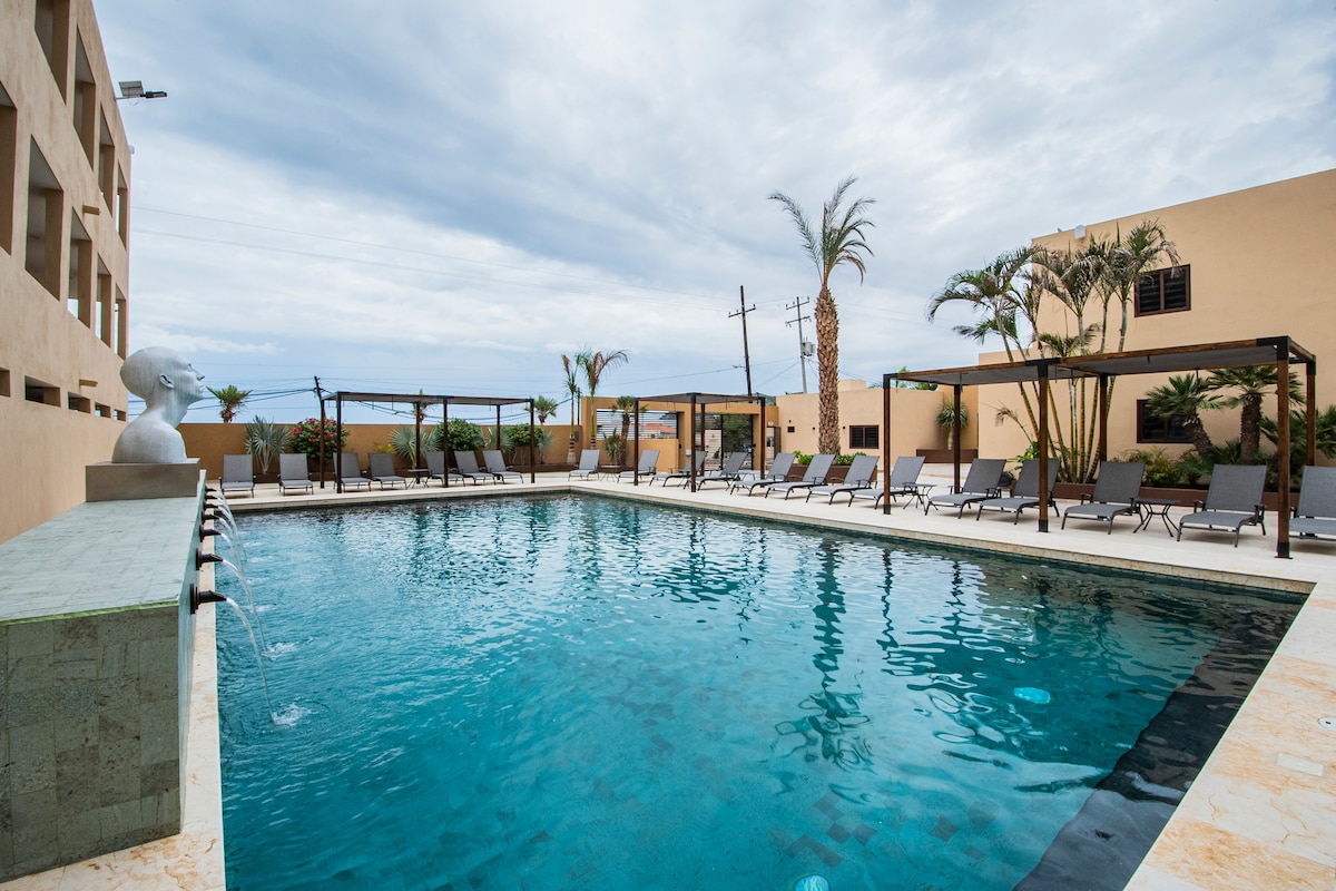 A spacious outdoor pool area is surrounded by relaxing lounge chairs under shaded pergolas. Palm trees are positioned along the perimeter, enhancing the tropical ambiance. The water reflects the cloudy sky, creating a calm atmosphere for leisure and relaxation.