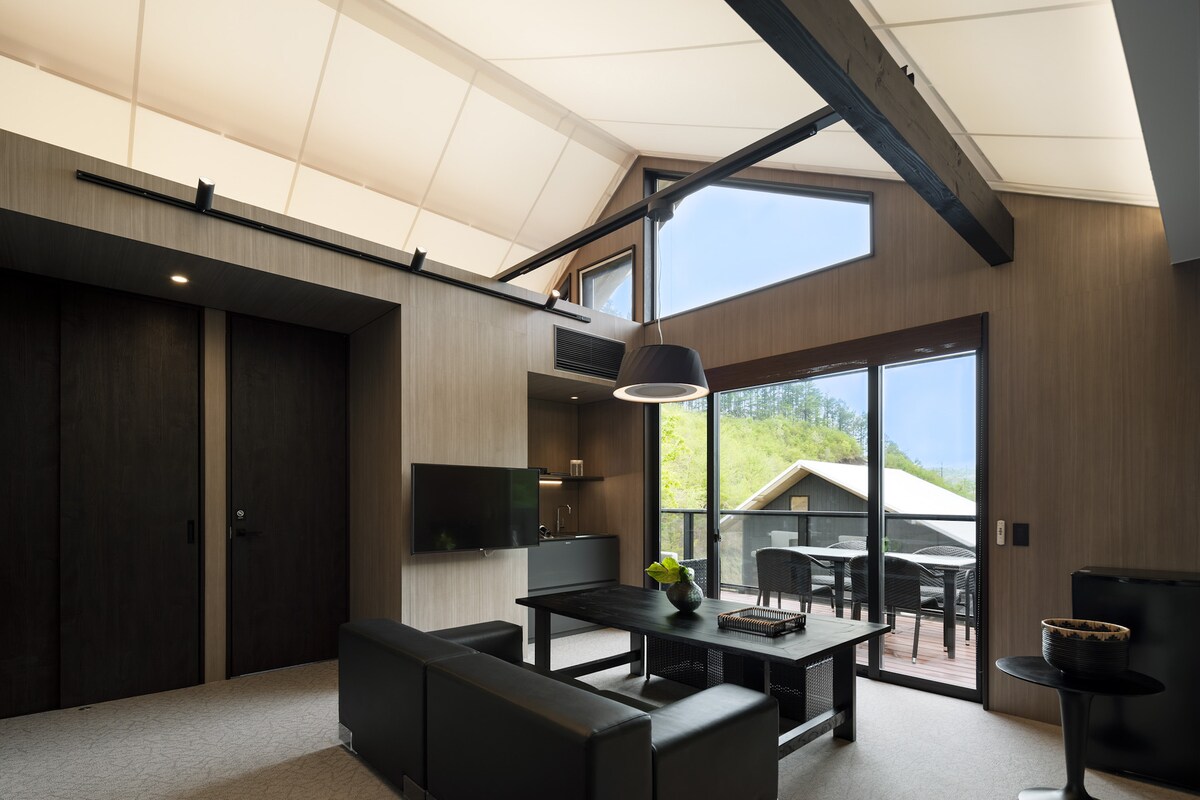 A modern living area features a dark leather sofa and a wooden dining table. Large windows allow natural light to illuminate the space, while a view of the outdoors is visible through the glass. Ceiling beams add architectural interest above.