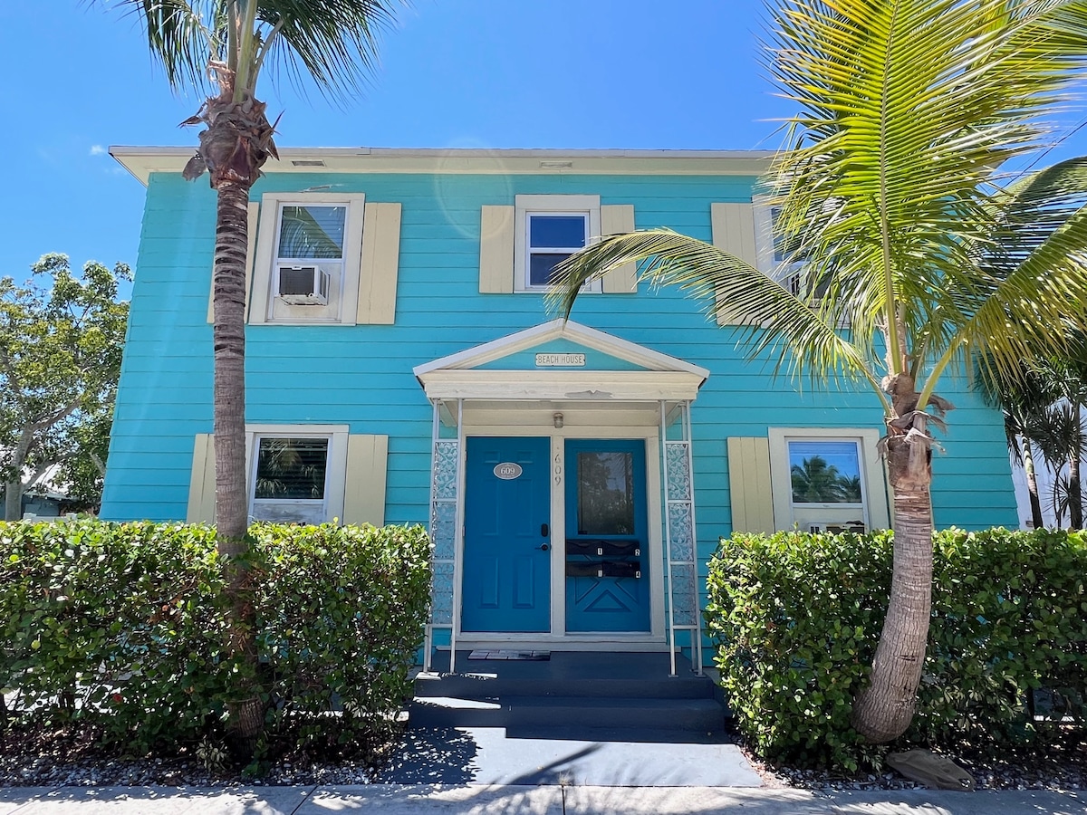 A vibrant blue exterior showcases a two-story building, framed by lush green foliage and palm trees. The entrance is highlighted by a welcoming blue door, flanked by light-colored shutters. Sunlight illuminates the bright facade, enhancing the cheerful atmosphere of the property.