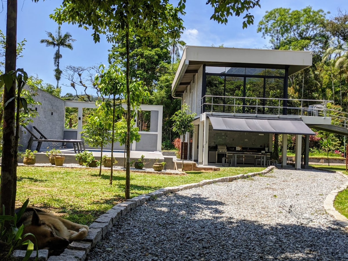 A modern structure stands amidst lush greenery. Large glass windows reflect the surrounding trees and natural light. A pathway of gravel leads through a landscaped garden with decorative pots, providing a serene approach to the property.