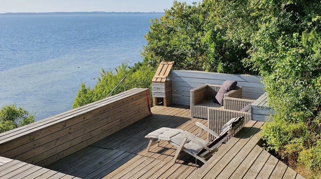 A spacious wooden terrace offers a panoramic view of the fjord, surrounded by greenery. Comfortable seating arrangements are provided with a pair of lounge chairs and a small table. Natural light shines brightly, enhancing the serene outdoor atmosphere.