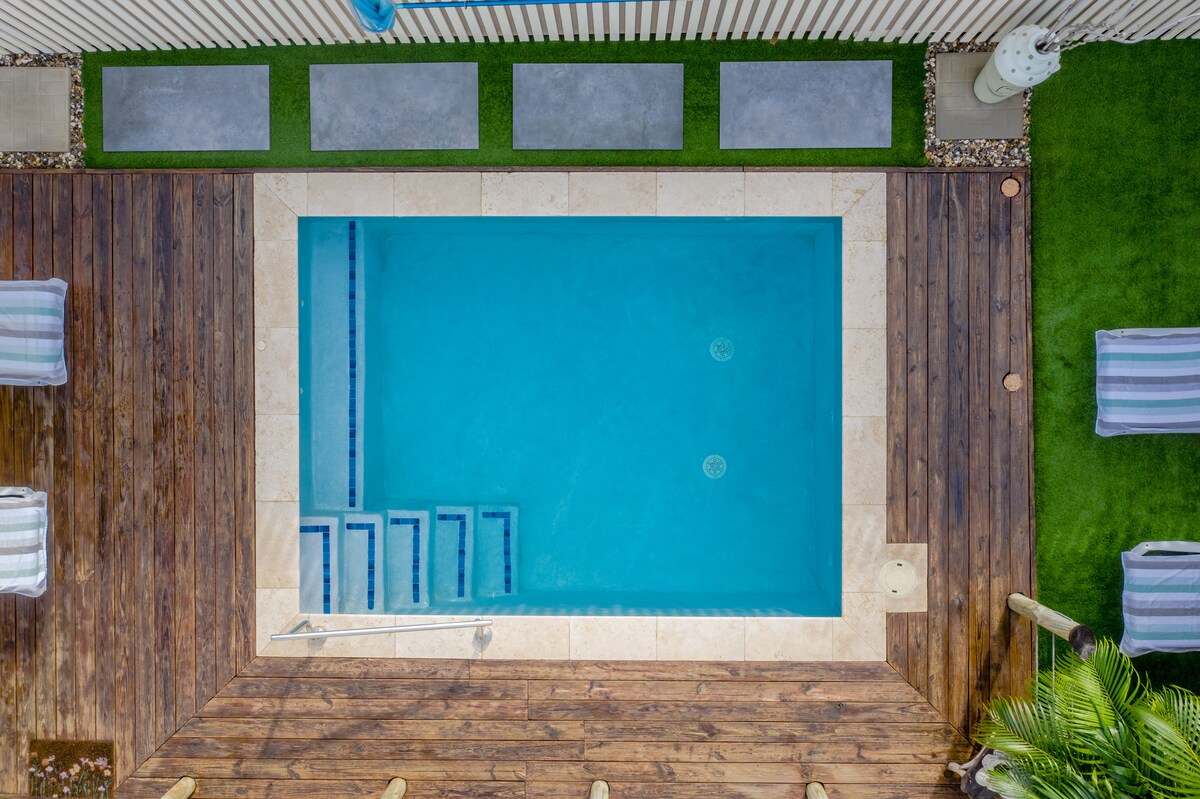 An aerial view of a private outdoor pool highlights its clear blue water, set within a rectangular stone border. Surrounding the pool are light-colored stone tiles and wooden decking, with lounge chairs arranged nearby, providing a relaxing space for sunbathing.