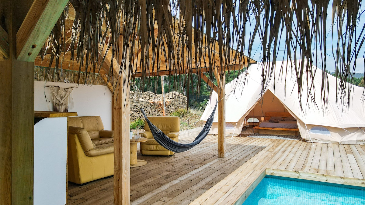 An outdoor terrace is shaded by a thatched roof, featuring a cozy seating area with a light-colored sofa and a hammock. A spacious tent is positioned nearby, while a small swimming pool adds a refreshing element to the space.