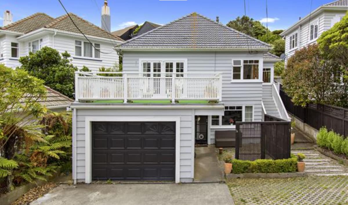 A spacious character house is presented with a light grey exterior and a prominent front deck. The garage door is visible, along with well-maintained landscaping featuring greenery and decorative pots. The surrounding neighborhood is highlighted by residential greenery, contributing to a peaceful setting.