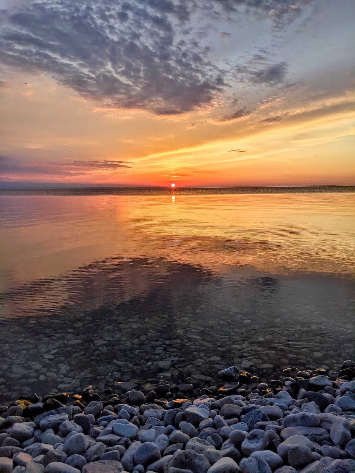 A tranquil shoreline features smooth stones lining the beach, reflecting soft colors of a sunset over calm waters. The sun sets on the horizon, casting warm hues that blend with the sky's clouds, creating a serene atmosphere along the beach.