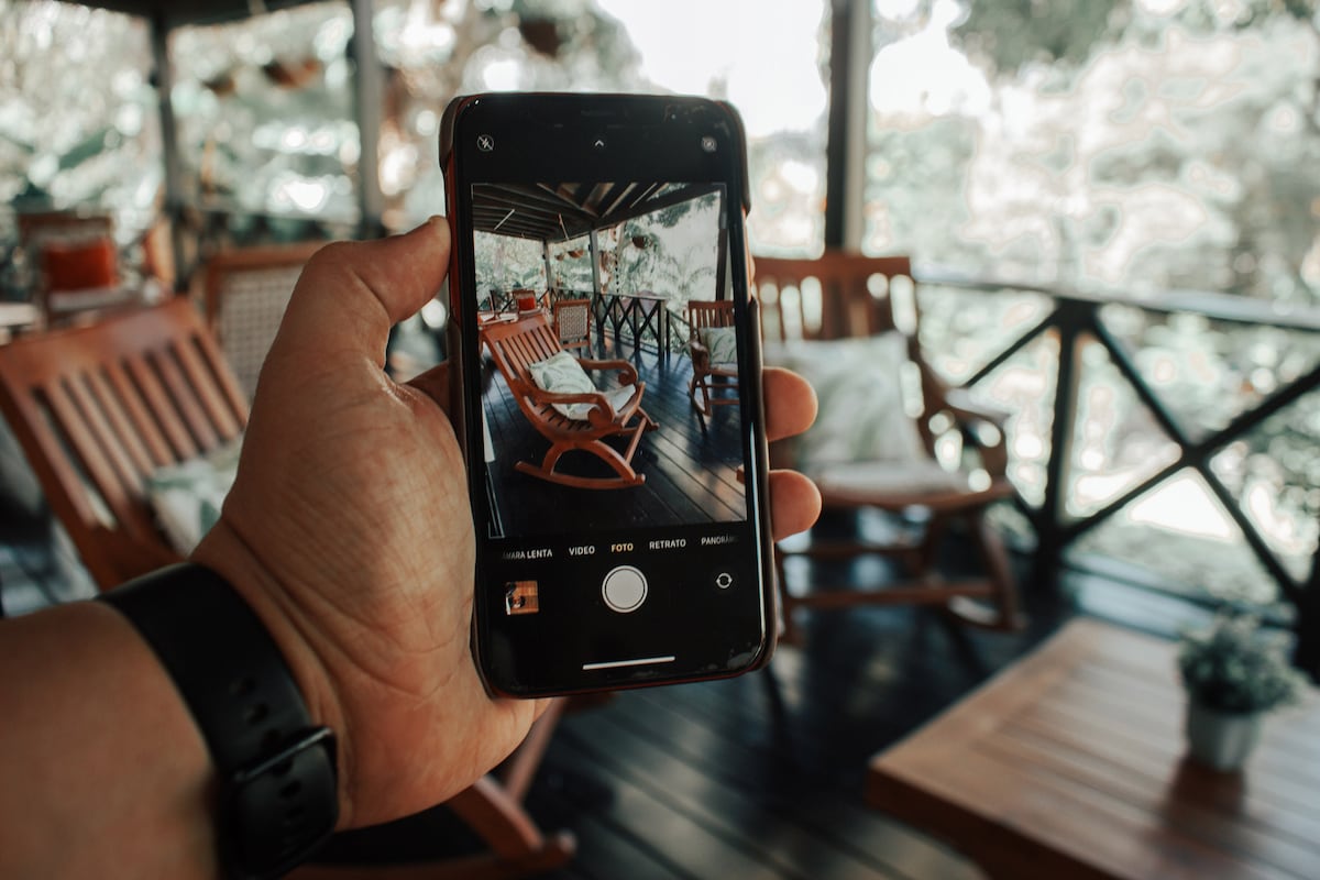 A hand holds a smartphone displaying a cozy seating area with wooden rocking chairs under a covered porch. Lush greenery is visible in the background, and natural light filters through, enhancing the inviting atmosphere of this outdoor space.