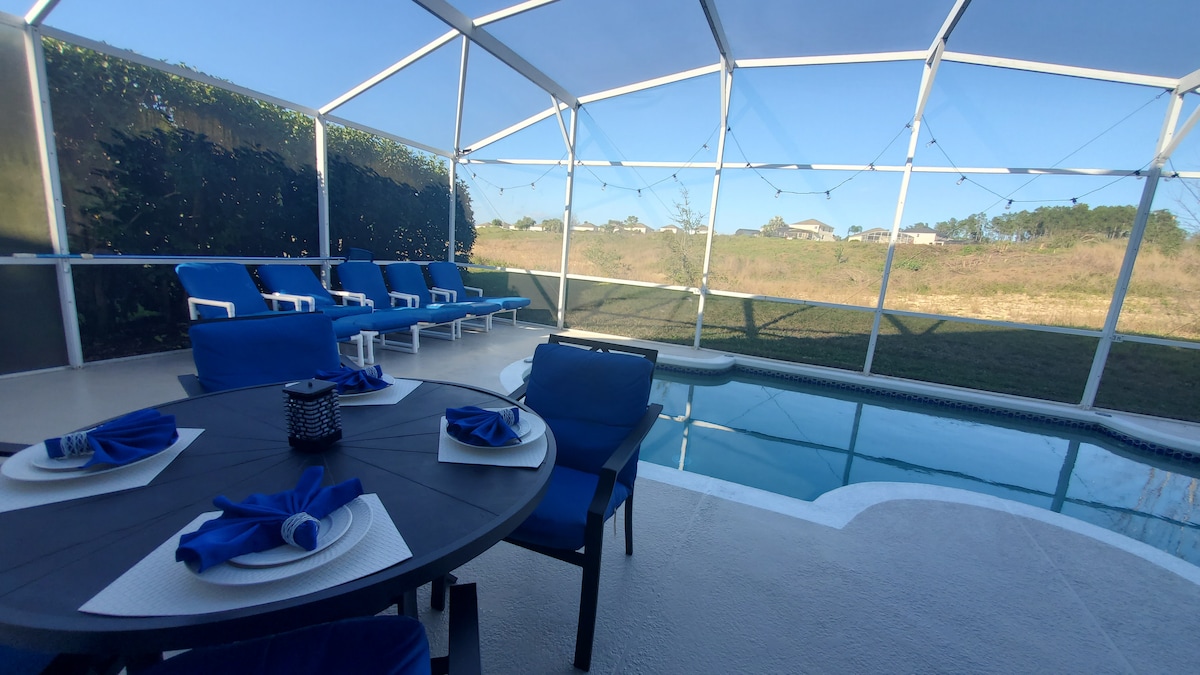 The outdoor pool area features a screened enclosure with views of the golf course beyond. A circular table is set with blue napkins and dinnerware, surrounded by blue chairs. Six lounge chairs are positioned alongside the shimmering pool, providing a comfortable space to relax.