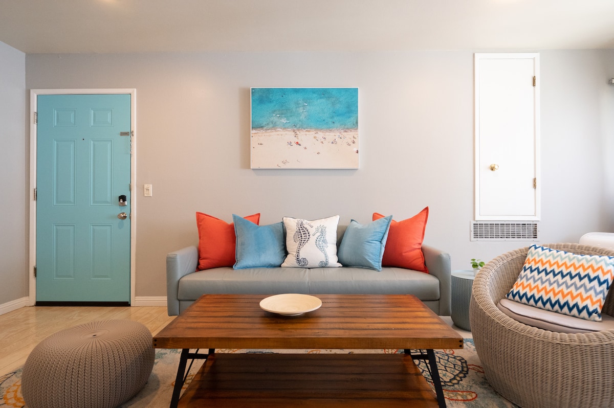 A modern living area features an Italian leather couch adorned with orange and blue accent pillows. A wooden coffee table sits in front, complemented by a circular pouf. The blue-toned artwork above the couch adds a pop of color, while the doorframe showcases a vibrant turquoise entrance.