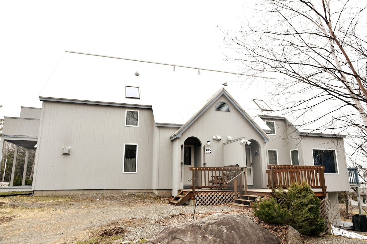 An exterior view of a two-story condo displays a symmetrical design with a sloped roof. A wooden deck with steps leads to the entrance, surrounded by gravel and a few shrubs. Large windows are present on either side, allowing natural light to enter.