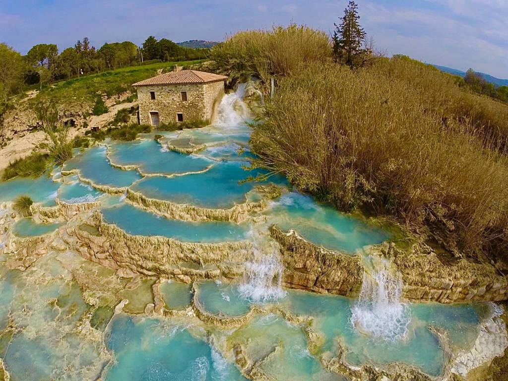 The image captures a natural landscape featuring cascading turquoise thermal waters flowing over terraced rock formations. A rustic stone building sits in the background, surrounded by lush greenery and trees, enhancing the serene ambiance of the outdoor setting.
