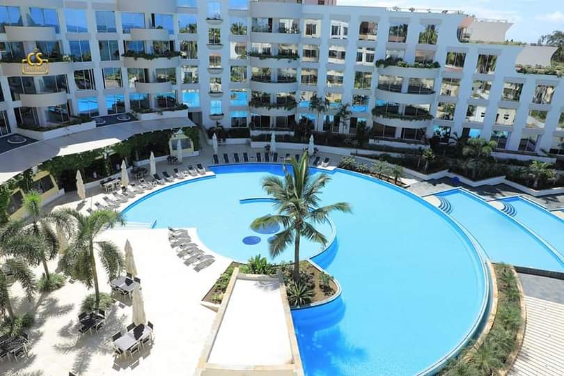 A sweeping view showcases a large, circular swimming pool surrounded by landscaped greenery and modern lounge chairs. The pool's calm waters reflect the surrounding architecture of the apartments, which are designed with multiple balconies overlooking this inviting outdoor space.